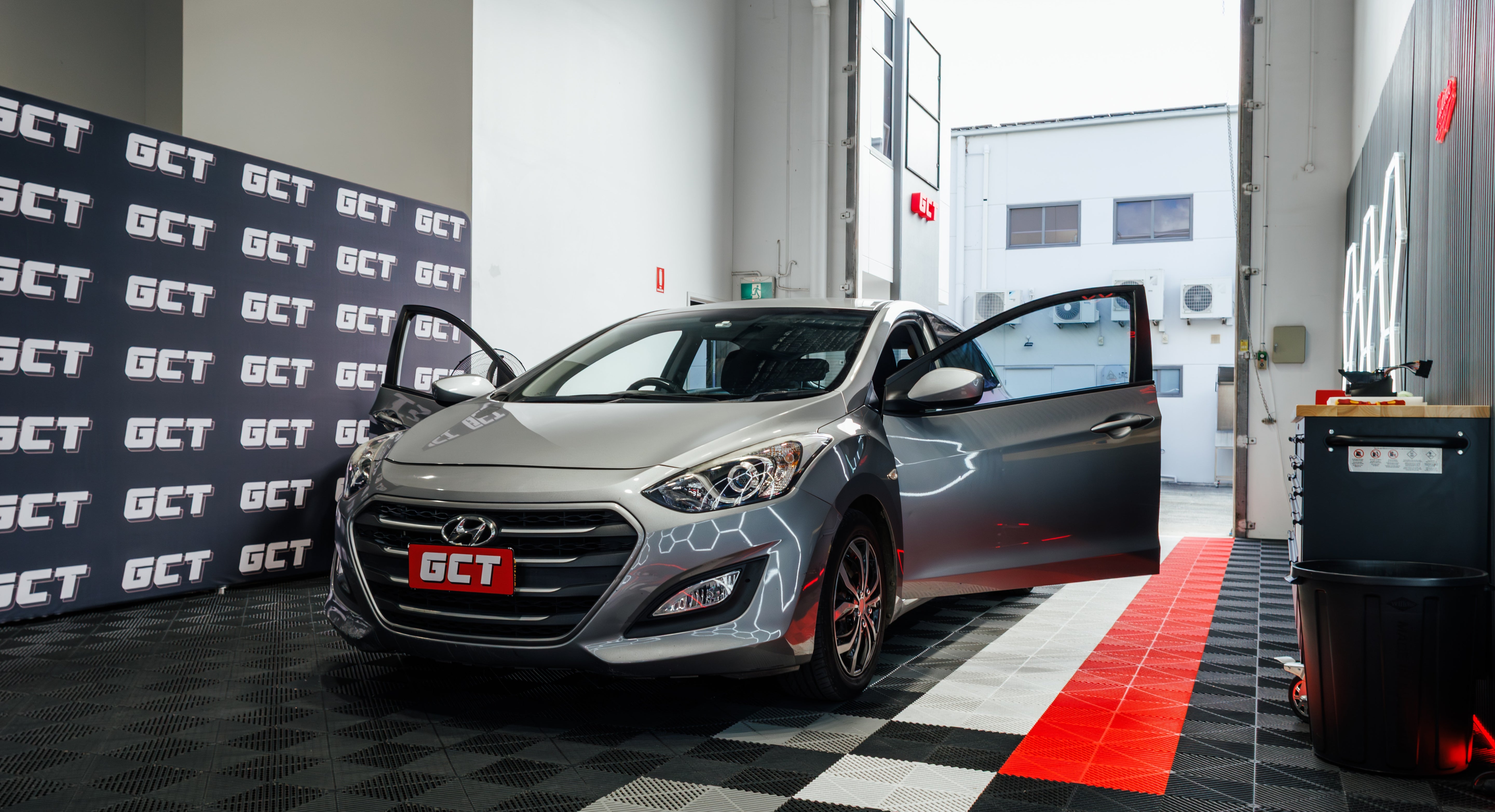 A silver Hyundai i30 with its front and side doors open, parked inside a garage or workshop. The car has a red GCT license plate and GCT branding on the bumper and side.