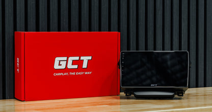 A GCT car head unit display screen next to its red retail packaging box on a wooden surface against a dark slatted background.
