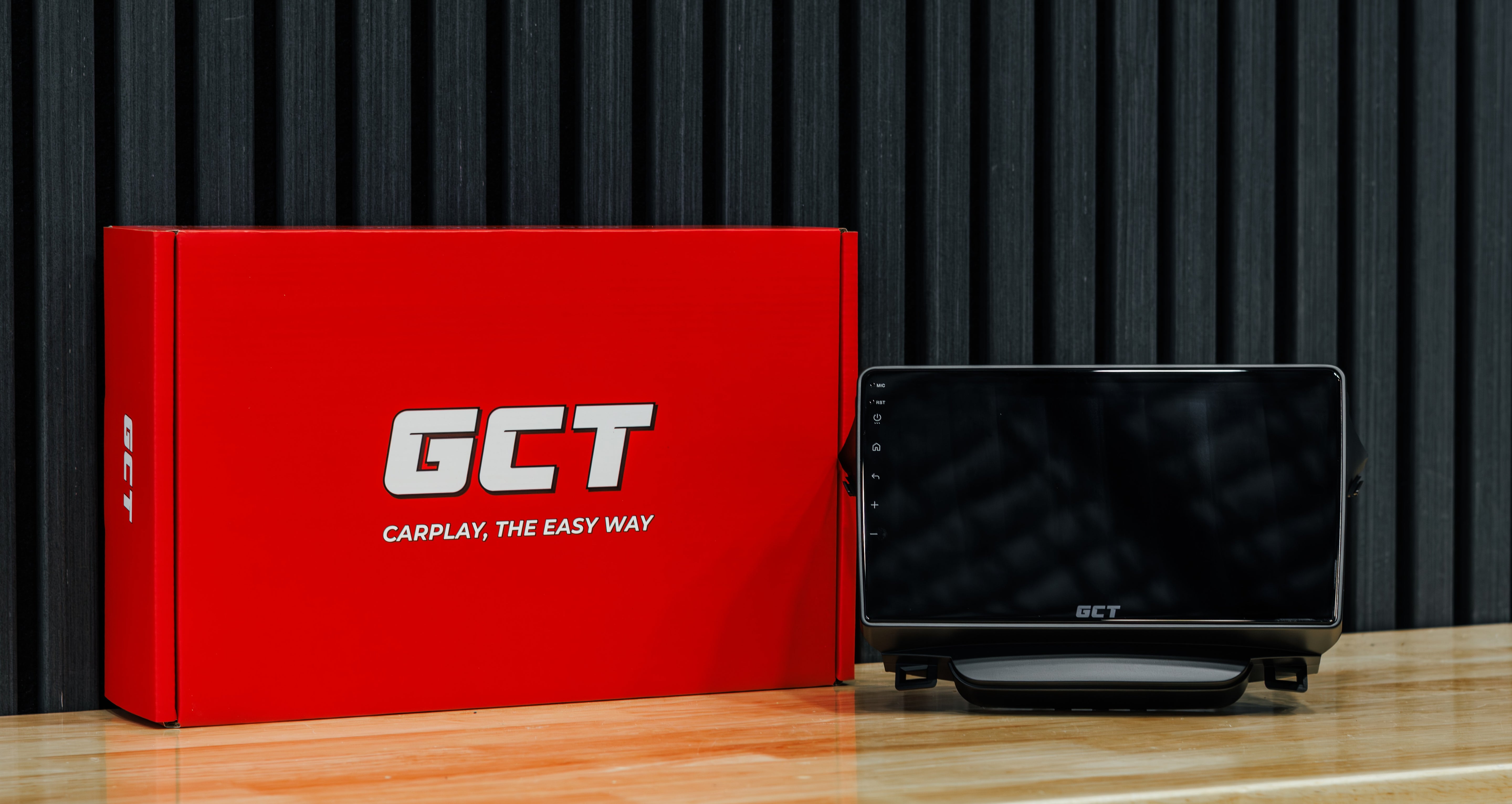 A GCT car head unit display screen next to its red retail packaging box on a wooden surface against a dark slatted background.
