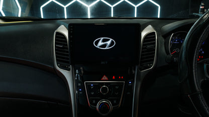 Close-up of a 2015 A 2015 Hyundai i30 dashboard with a large central touchscreen displaying the Hyundai logo, flanked by air vents and control buttons, with part of the steering wheel visible on the right.